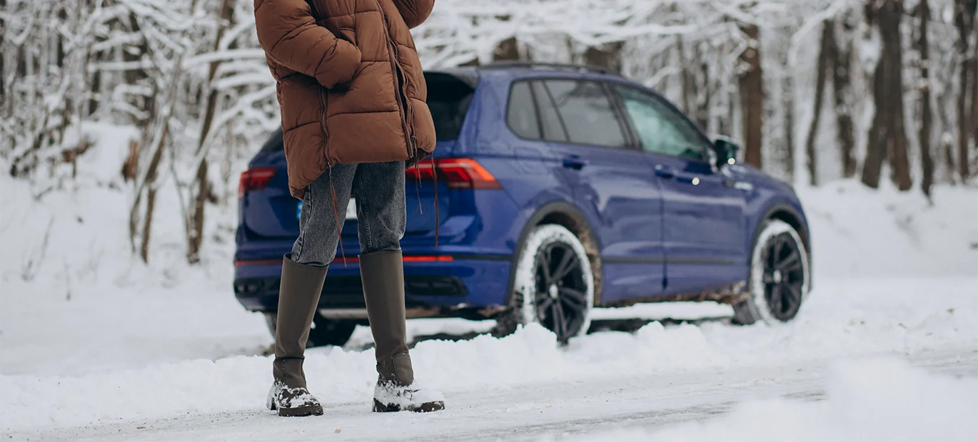 A woman stands beside her car, stranded on an icy road, stuck in the snow.