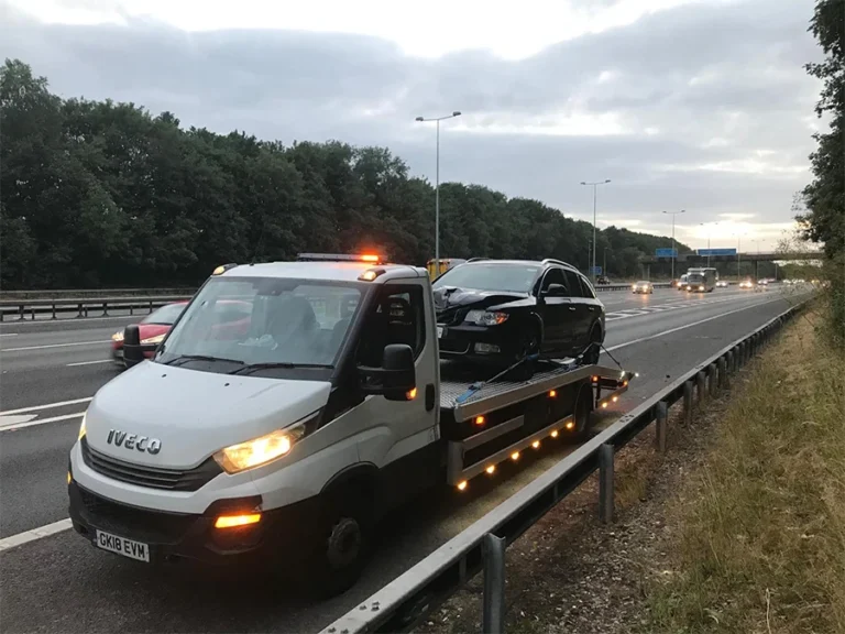 Professional vehicle recovery in Wembley: White truck towing a damaged black car on a main road.