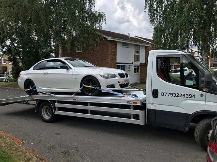 A white BMW coupe secured with professional straps on a white flatbed recovery truck during a car towing service in a residential area.