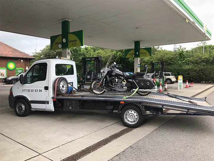 A black cruiser motorcycle securely loaded onto a white recovery flatbed truck for motorcycle towing at a BP petrol station.