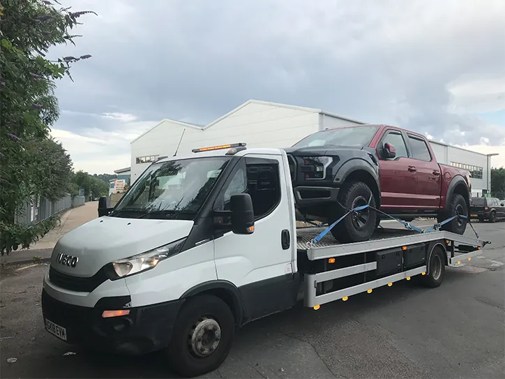 A white Iveco recovery truck providing 4x4 towing for a large red Ford Raptor pickup truck on a flatbed trailer.