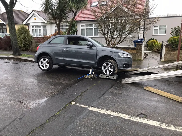A grey Audi hatchback being lifted by a trolley jack and placed on wheel skates for roadside assistance in a residential neighborhood.