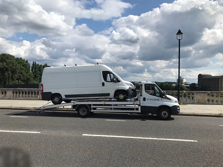 A white recovery flatbed truck performing a van towing service for a large white delivery van on a city bridge under a cloudy sky.