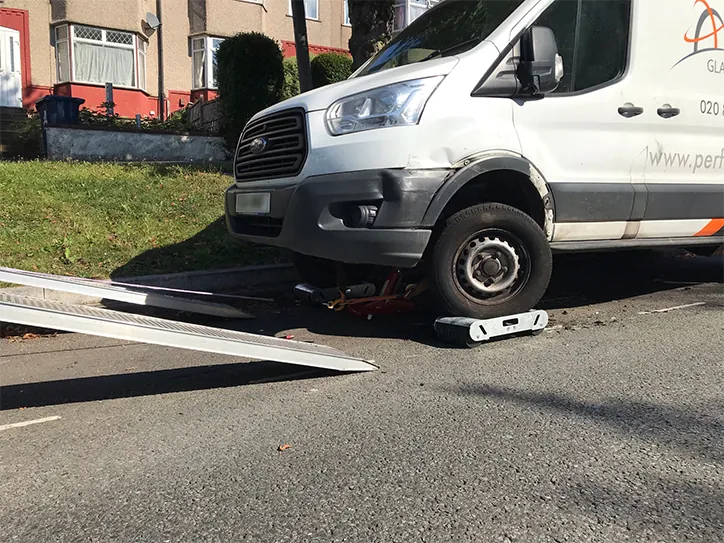 A damaged white Ford Transit van being prepared for vehicle recovery using wheel skates and a jack to load it onto a flatbed ramp in a residential street.